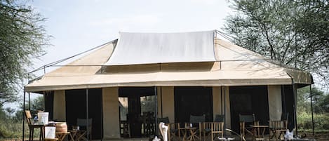 A safari tent with a large canvas roof, surrounded by wooden furniture and a small table.