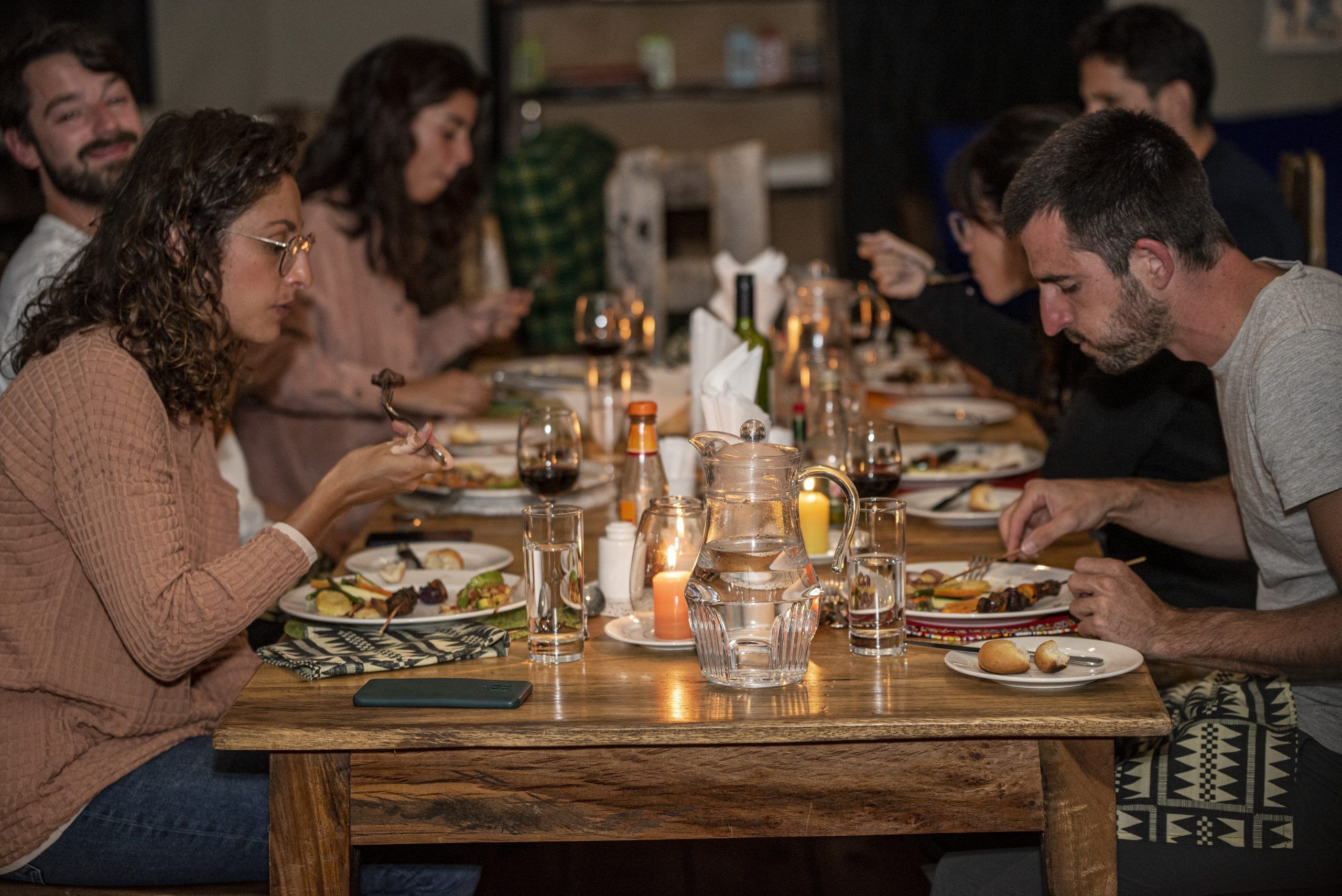 A group of people dining at a wooden table with various dishes, glasses, and a candle.