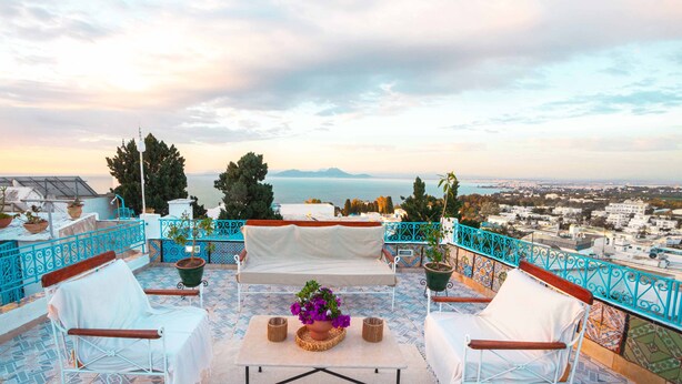 Una terraza con sillas y mesa de metal blanco, una planta en maceta y vistas al mar y al paisaje urbano.