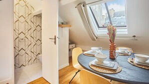 Un coin repas avec une table ronde dressée pour quatre personnes, une fenêtre donnant sur des immeubles et un mur orné d’un motif géométrique.