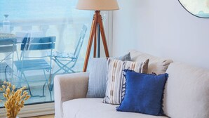 A living room with a sofa, a lamp, a round table, and a view of the sea through a large window. - La Vague Bleue – water feet S 04 (Marsa)