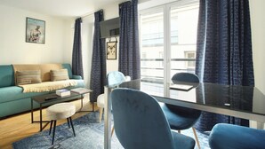 Een moderne woonkamer met een bank, een glazen tafel en stoelen. Er is een groot raam met gordijnen, een flatscreen televisie en een ingelijst kunstwerk aan de muur.