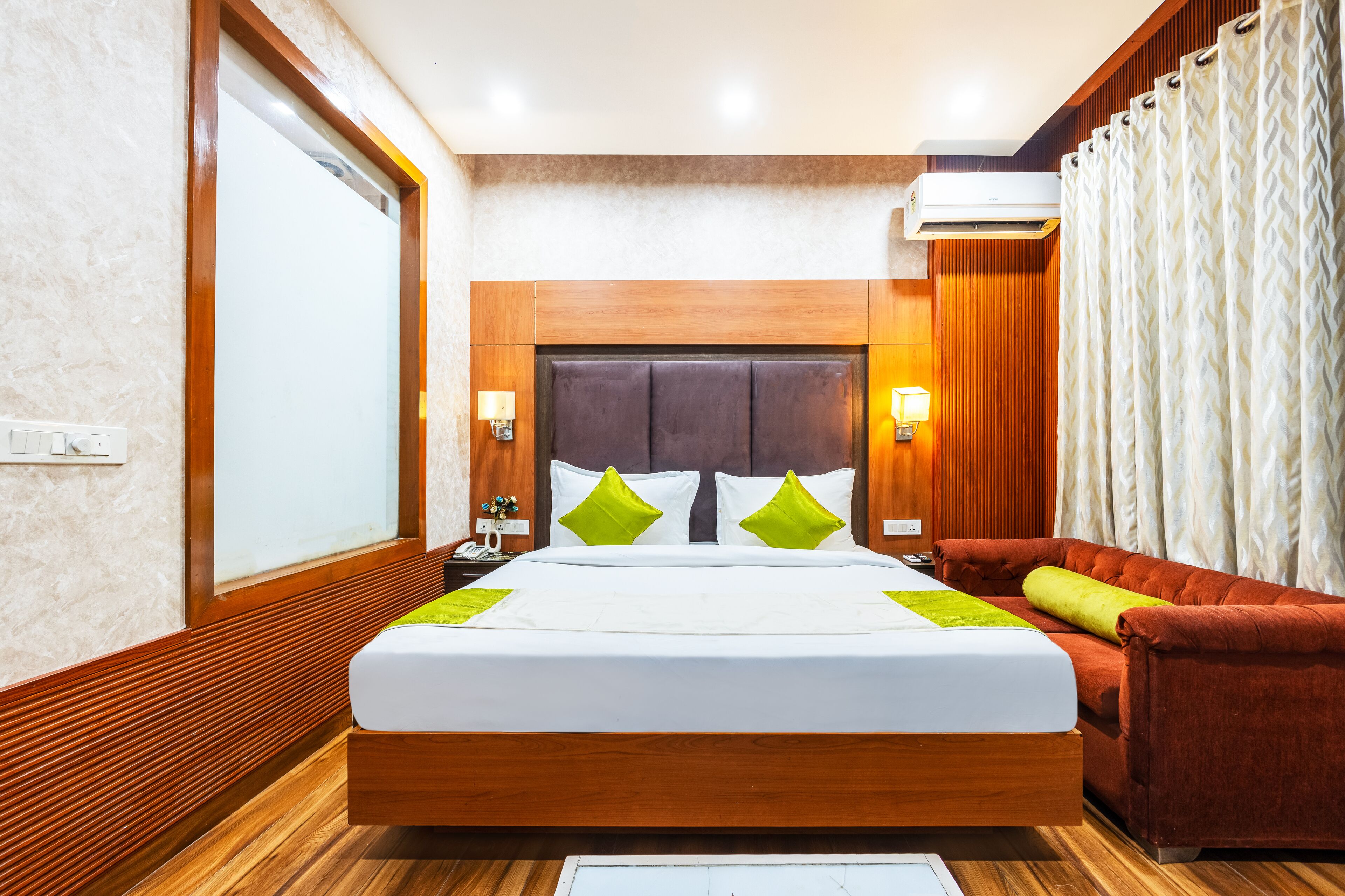 A hotel room with a large bed, wooden headboard, two bedside lamps, a red sofa, and a window with curtains.