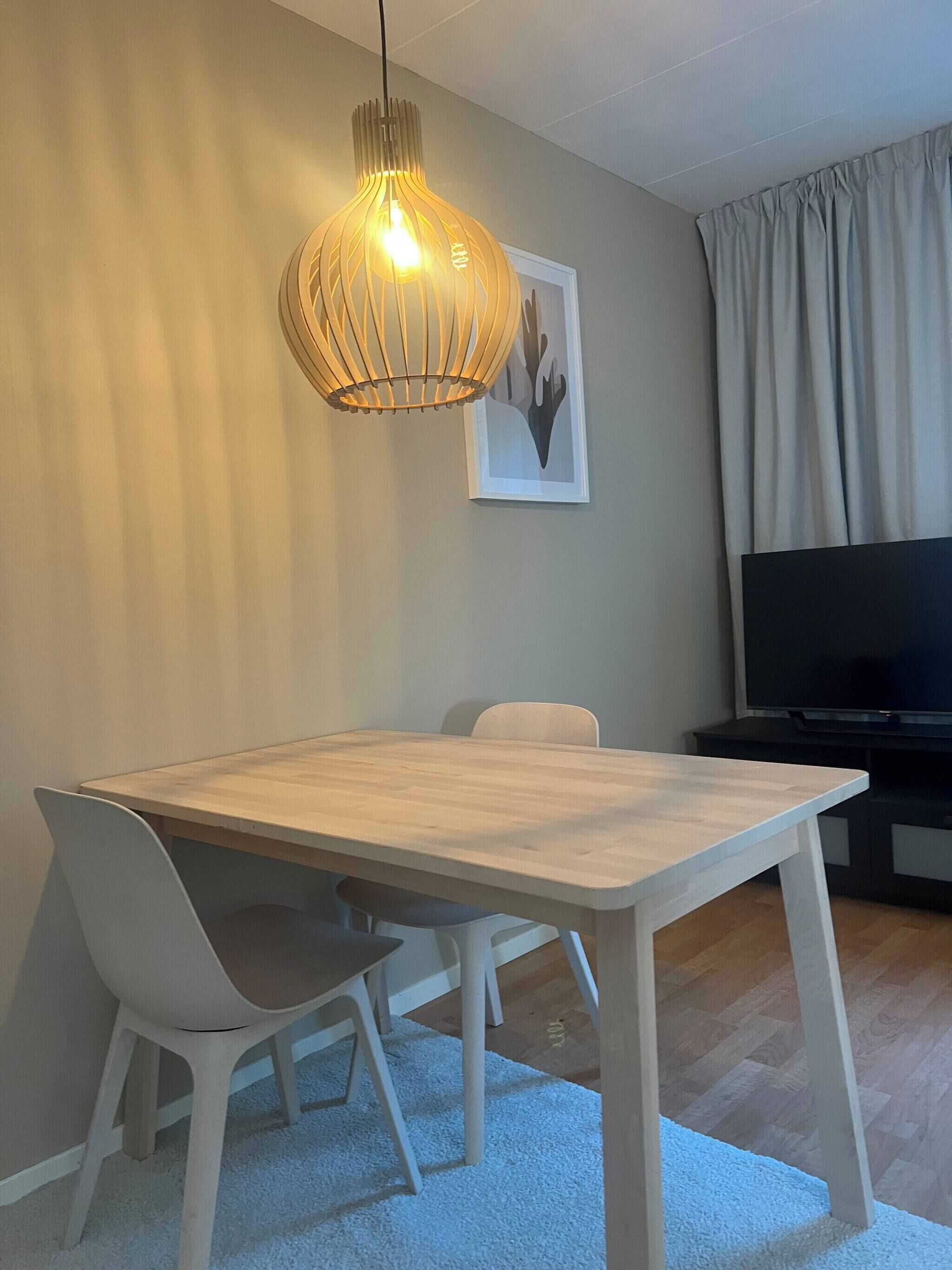 A dining area with a wooden table and white chairs, a hanging light fixture, a framed artwork on the wall, and a TV on a stand.