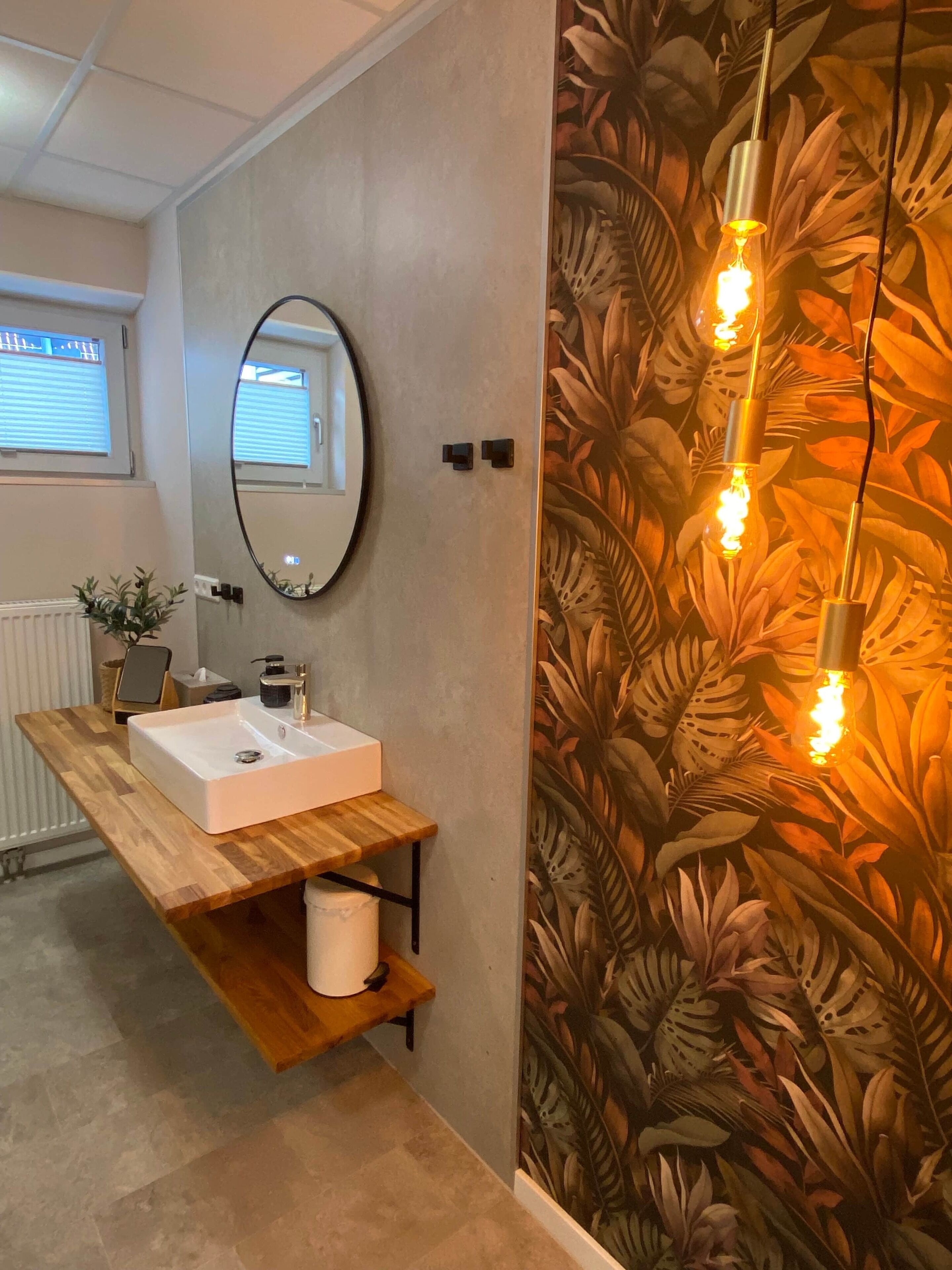Un baño con tocador de madera, lavamanos blanco, espejo y pared con estampado de hojas tropicales.