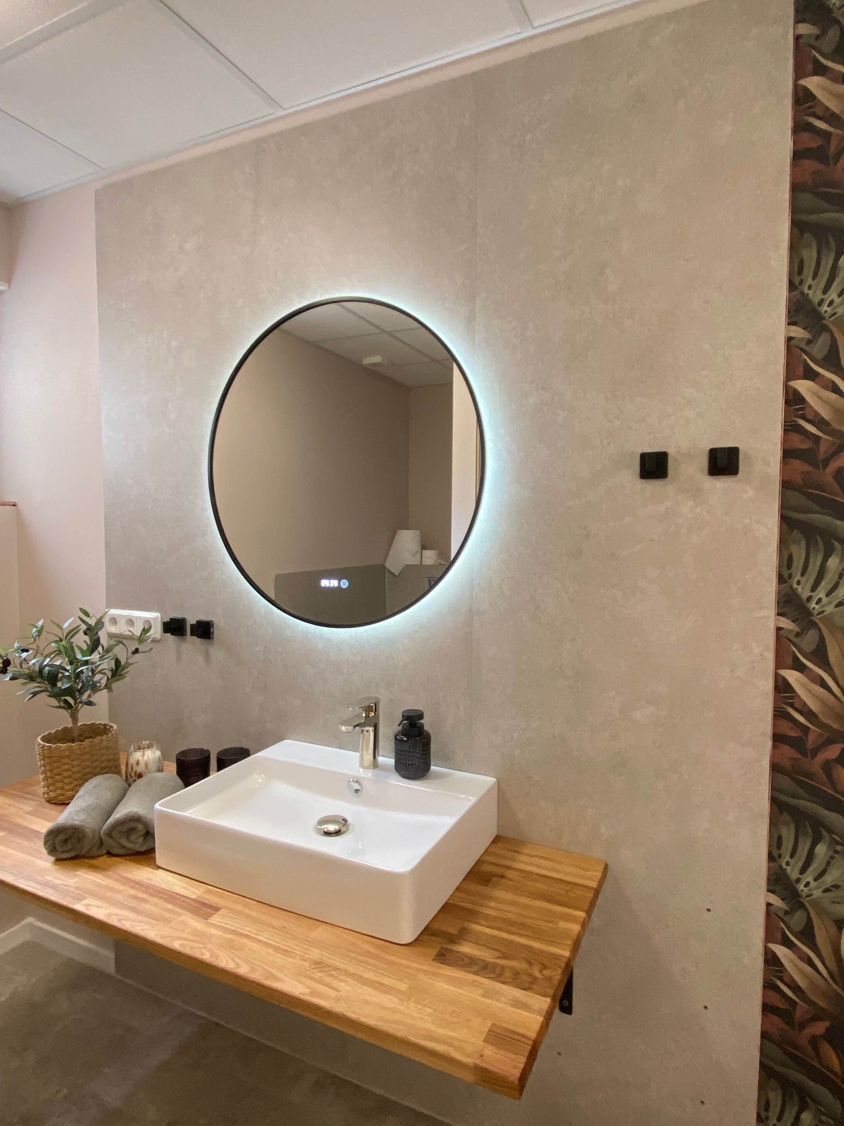 Une salle de bain avec un lavabo blanc, un miroir rond avec un éclairage intégré et un meuble-lavabo en bois.