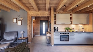 Une cuisine moderne avec des meubles gris, un four encastré et un évier. Le plafond est en bois avec des poutres apparentes, il y a un canapé gris et une petite table avec une lampe.