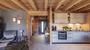 A modern kitchen with grey cabinets, a built-in oven, and a sink. There is a wooden ceiling with exposed beams, a grey sofa, and a small table with a lamp.