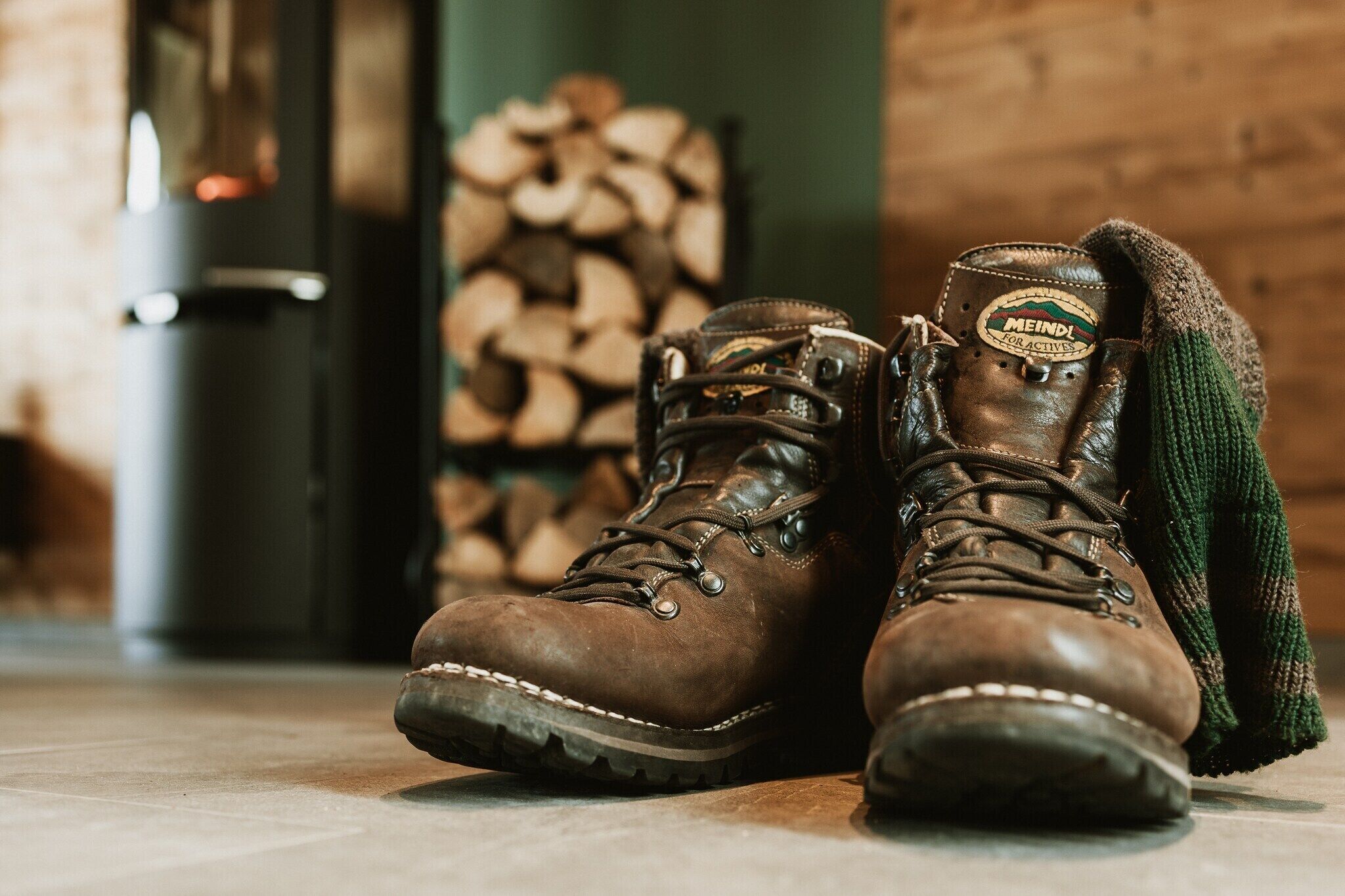 A pair of brown hiking boots with a logo on the tongue, placed on a tiled floor with a pile of firewood in the background.