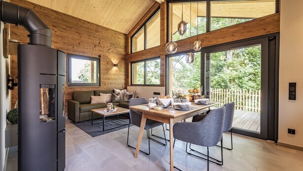 Een modern interieur met een houten plafond, een zwart fornuis, een eethoek met een tafel en stoelen, en grote ramen met uitzicht op groen.