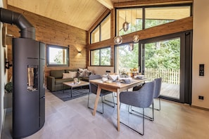 A modern interior with a wood-paneled ceiling, a black stove, a dining area with a table and chairs, and large windows offering a view of greenery. - Modernes ebenerdiges Chalet im Vital Camp Bayerbach (Bayerbach)