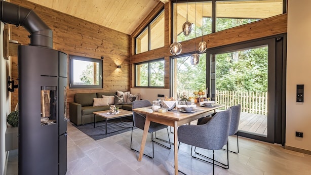 A modern interior with a wood-paneled ceiling, a black stove, a dining area with a table and chairs, and large windows offering a view of greenery. - Ebenerdiges gemütliches Chalet im Vital Camp Bayerbach (Bayerbach)