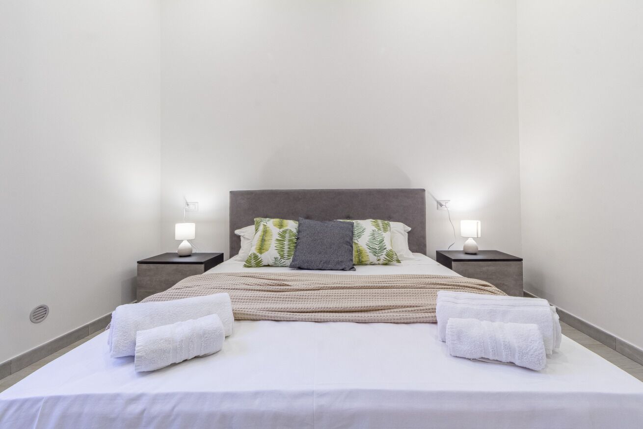 Een net opgemaakt bed met witte lakens en twee witte handdoeken, een grijs kussen en een deken met een patroon. Er staan twee nachtkastjes met lampen bij.
