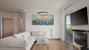 Ein modernes Wohnzimmer mit einer Eckcouch, einem Glastisch, einem großen abstrakten Gemälde, einer Deckenleuchte und einem Fernseher an der Wand.