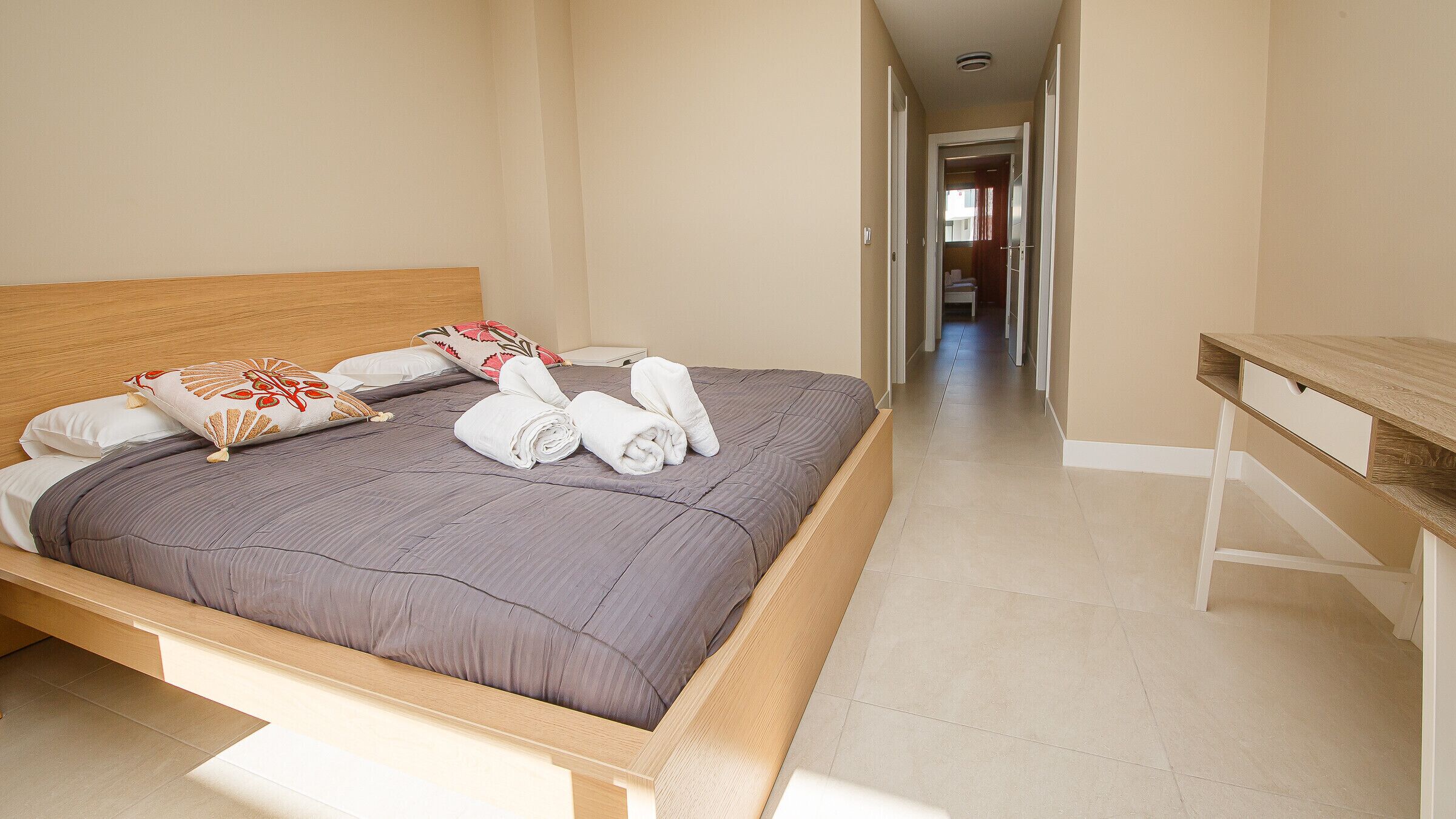 Un lit bien fait, avec une tête de lit en bois, une couvre-lit grise et des coussins décoratifs. Une table de chevet en bois, dotée d’un tiroir, se trouve à droite du lit. La pièce est dotée d’un sol carrelé et un couloir mène à une autre pièce.