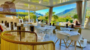 An outdoor dining area with tables and chairs, a wicker chair in the foreground, and a view of mountains in the background. - SCHLOSSHOF SÜDTIROL – Hotel & Camping Resort (Lana)