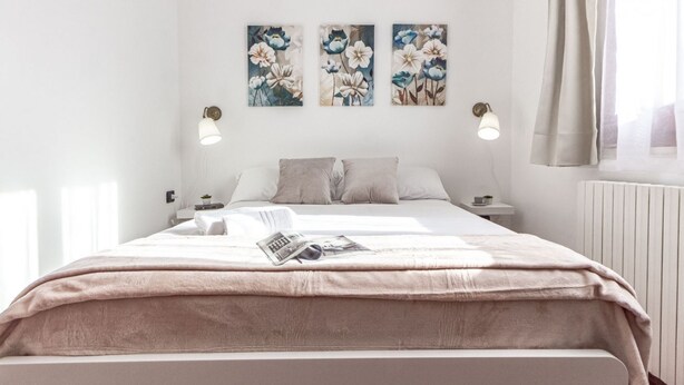 Un dormitorio moderno con una cama grande, una mesita de noche y un velador. Hay tres lámparas fijadas a la pared y una ventana con cortinas.