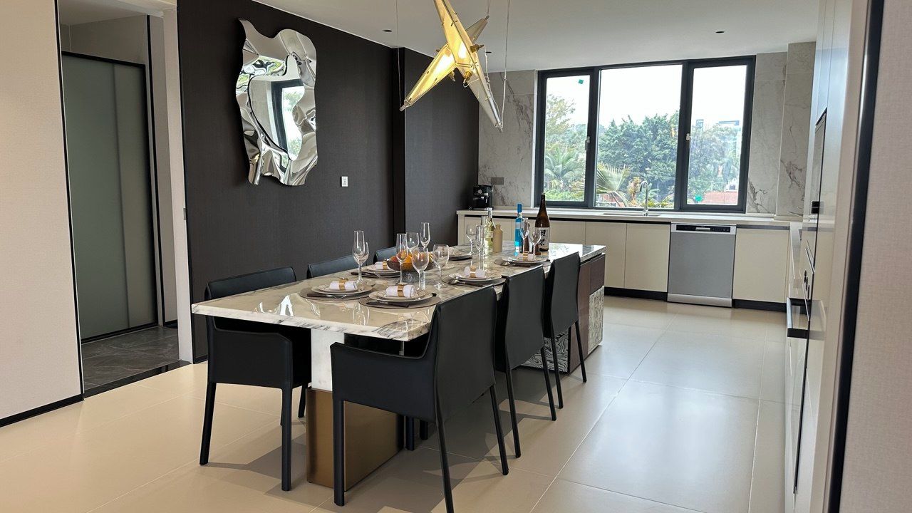 A modern dining area with a large table set for six, a kitchen in the background, and a large window providing natural light.