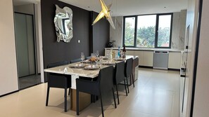 Un comedor moderno con una mesa grande dispuesta para seis personas, una cocina al fondo y un amplio ventanal que proporciona luz natural.