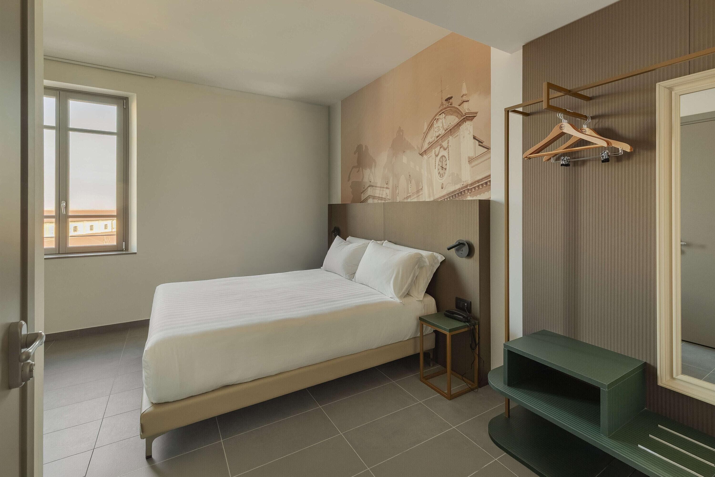 Quarto de hotel com cama, uma pequena mesa, um espelho e um porta-roupas fixado na parede.