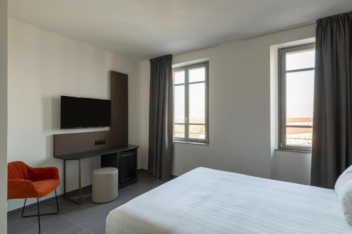 Een hotelkamer met een bed, een bureau met televisie, een stoel en twee ramen met gordijnen.