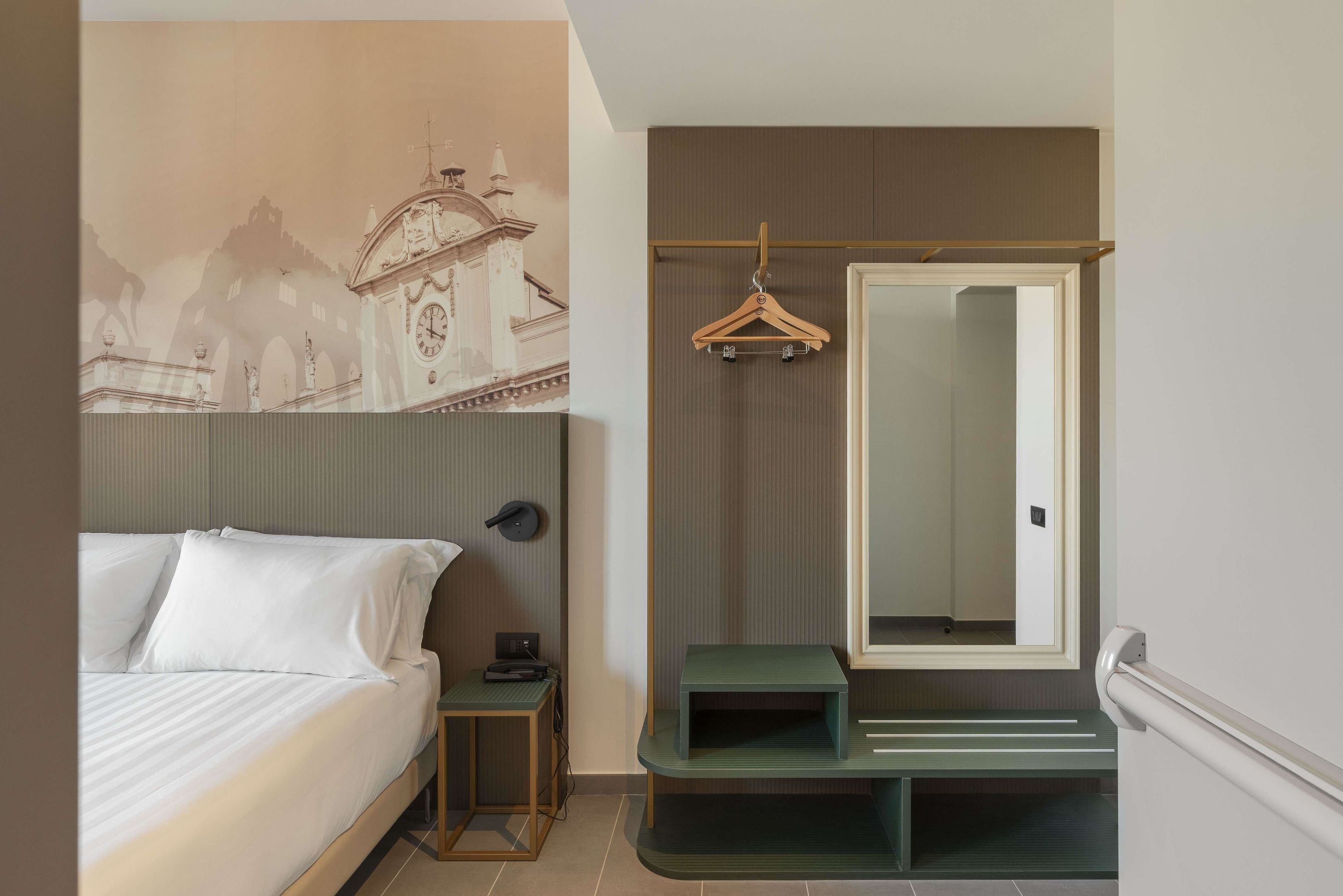 Una habitación de hotel con cama, mesita de noche, espejo y perchero en la pared.