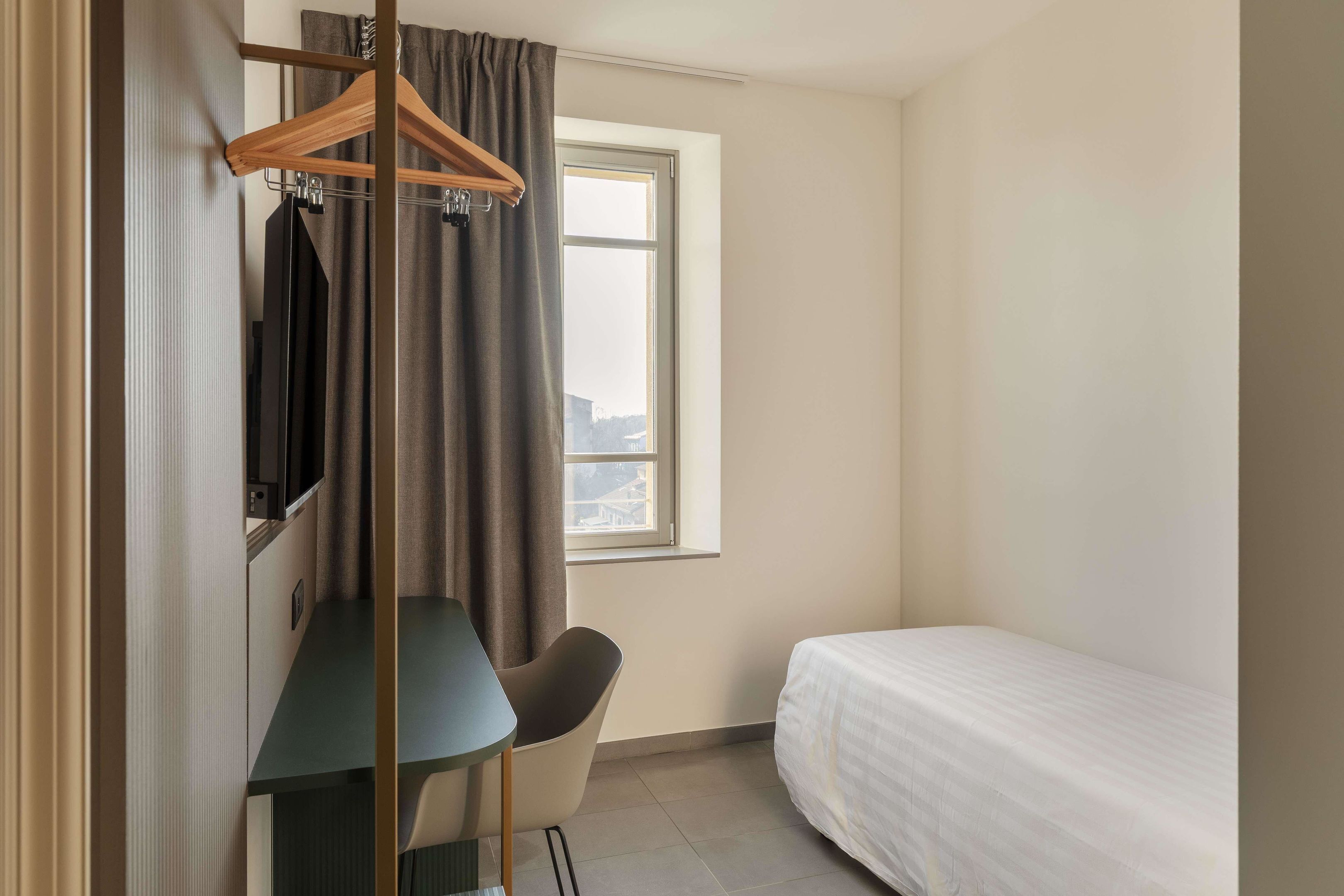 A small hotel room with a bed, a desk, a chair, a TV, and a window with curtains.