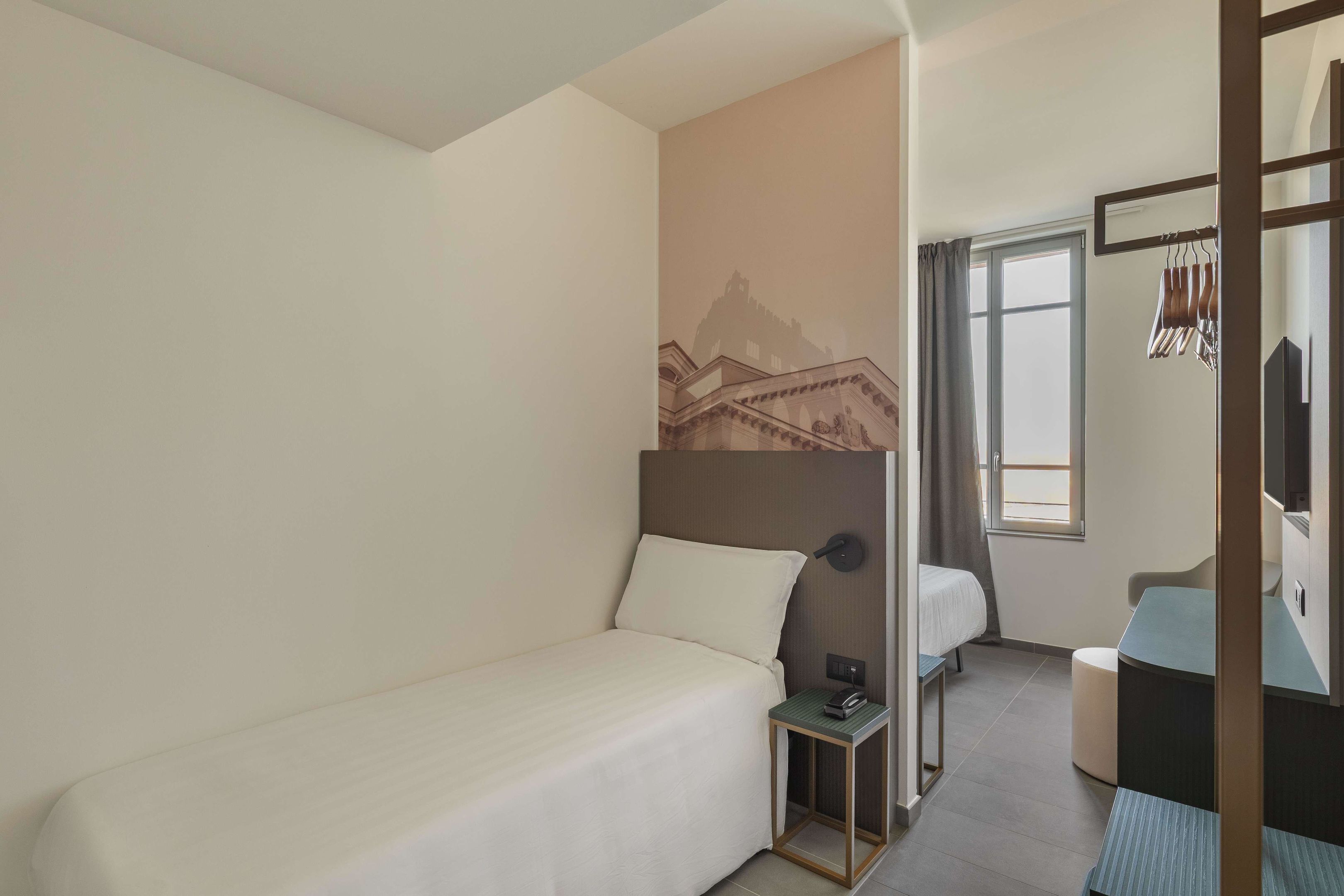 Ein kleines, ordentlich eingerichtetes Hotelzimmer mit einem Einzelbett, einem kleinen Tisch, einem Stuhl, einem Kleiderschrank und einem Fenster mit Vorhängen.