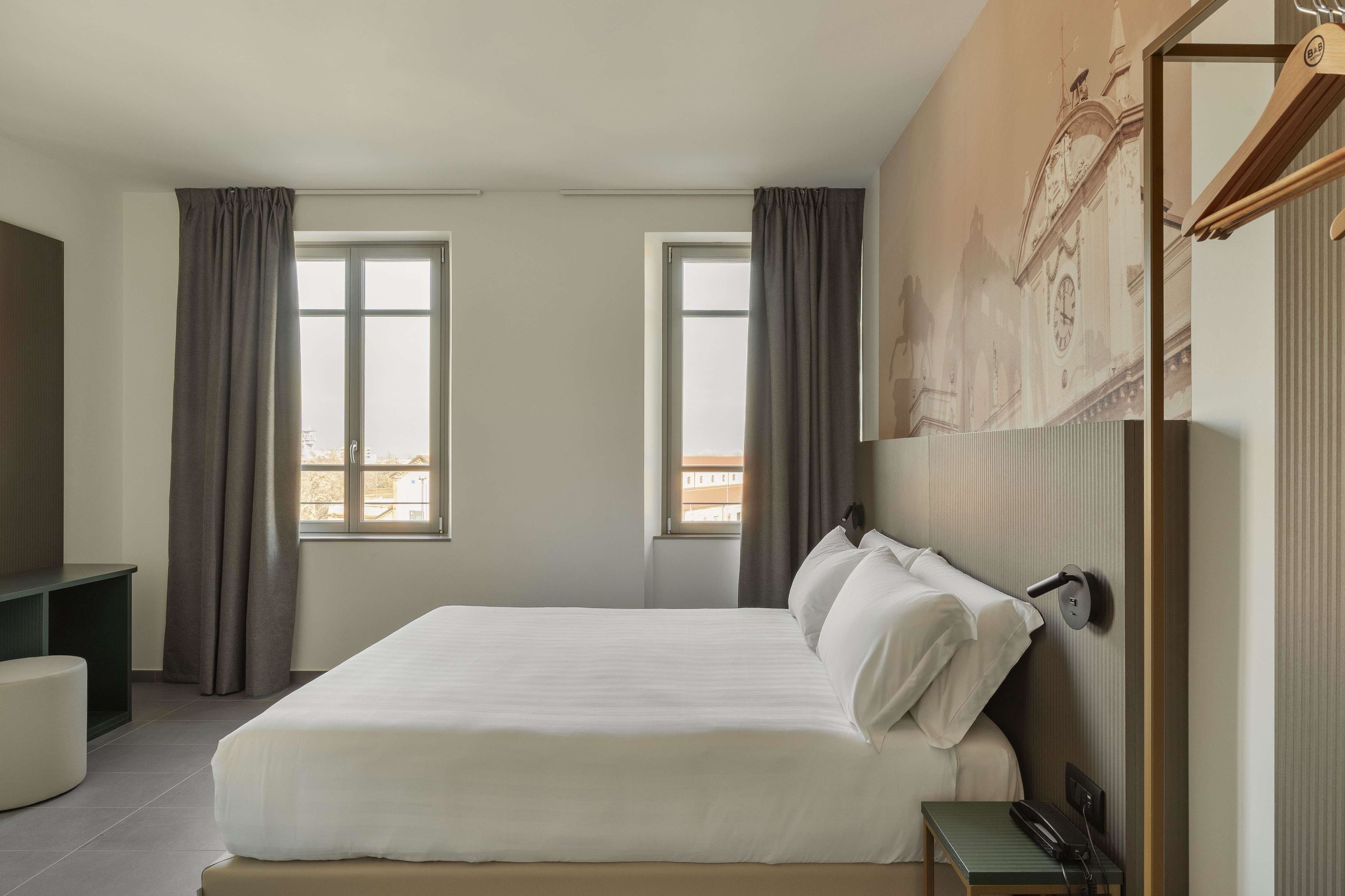 A hotel room with a large bed, two windows with curtains, a desk, and a wall-mounted telephone.