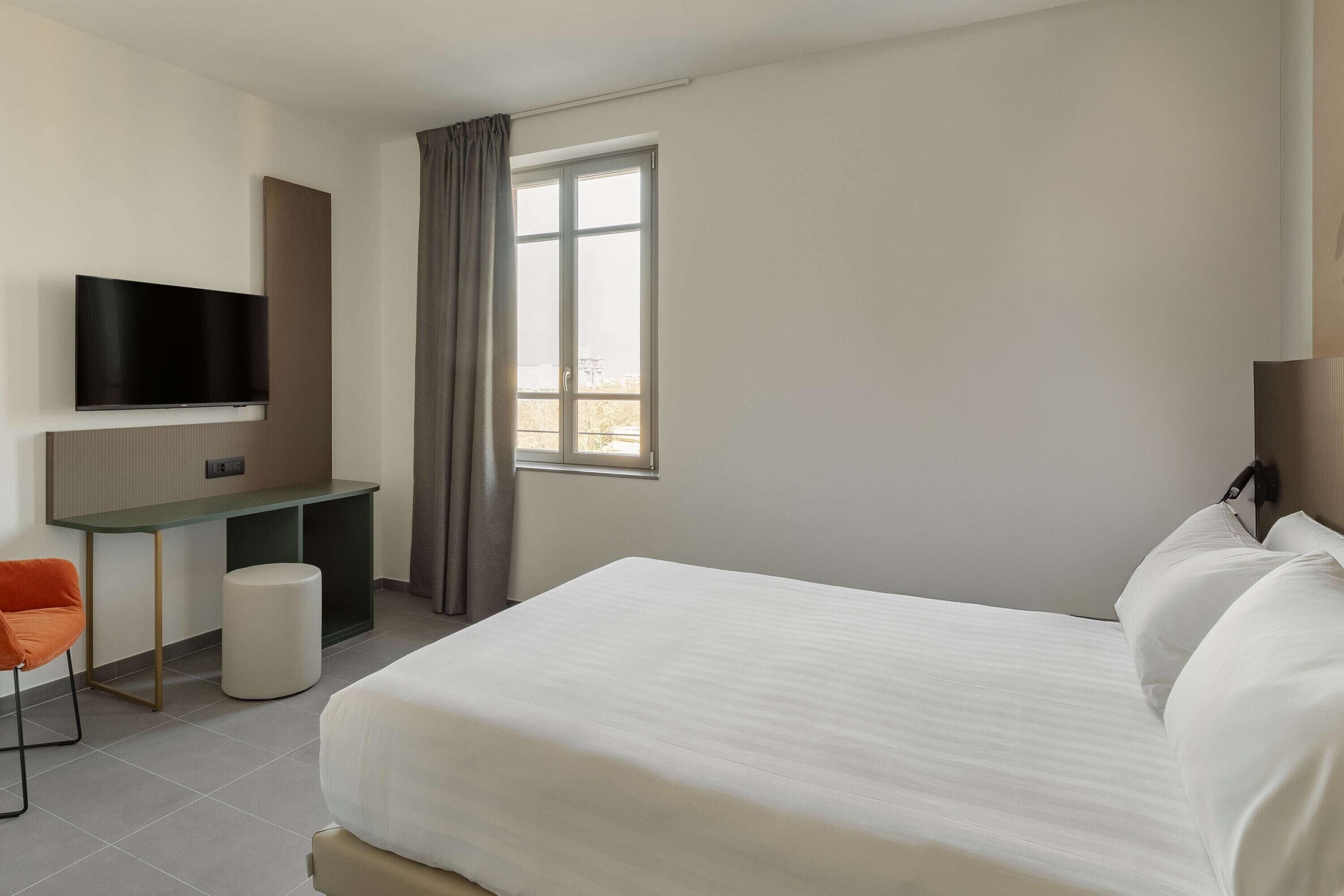 A hotel room with a bed, a desk, a chair, a television, and a window.
