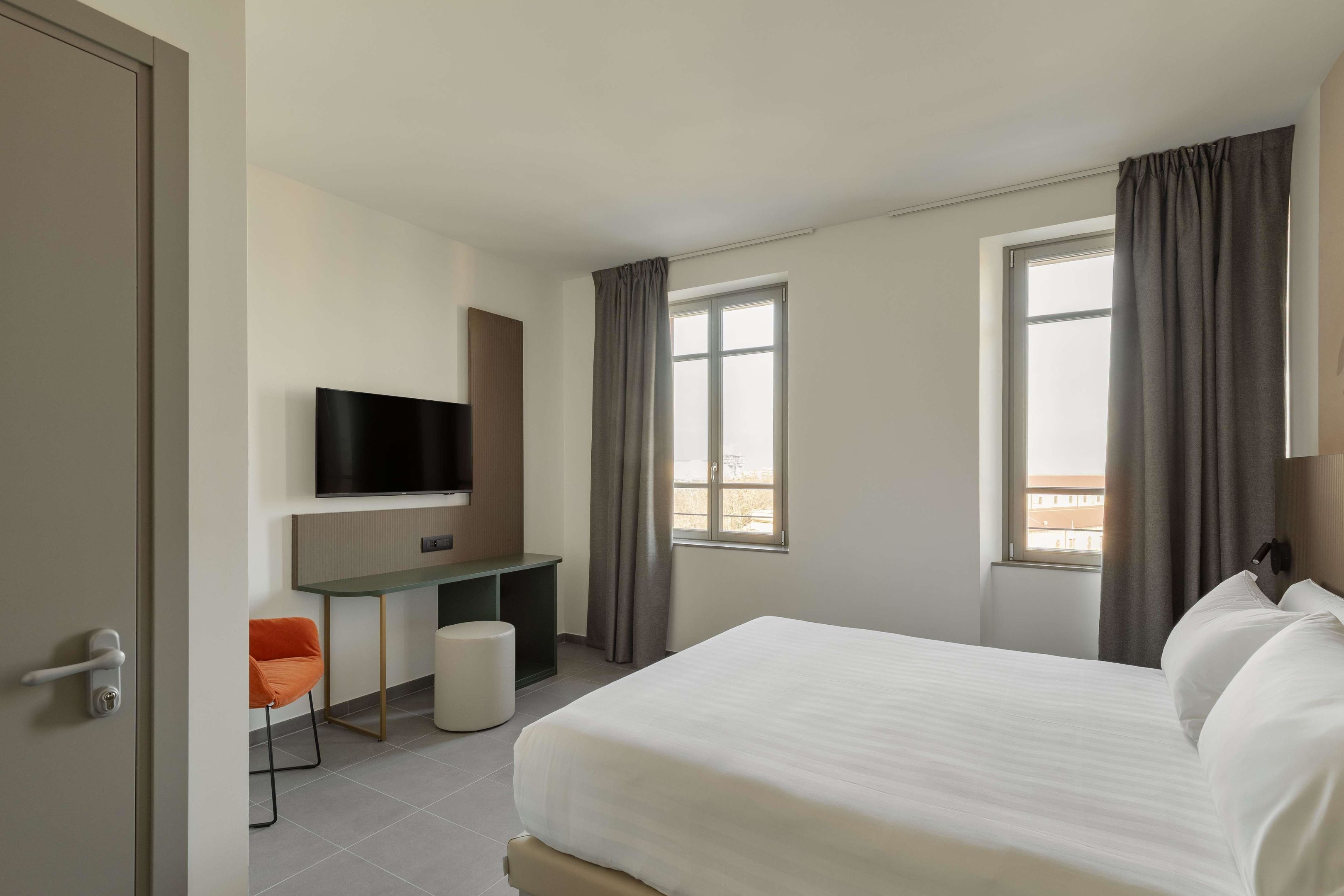 A hotel room with a bed, a desk, a chair, a television, and two windows with curtains.