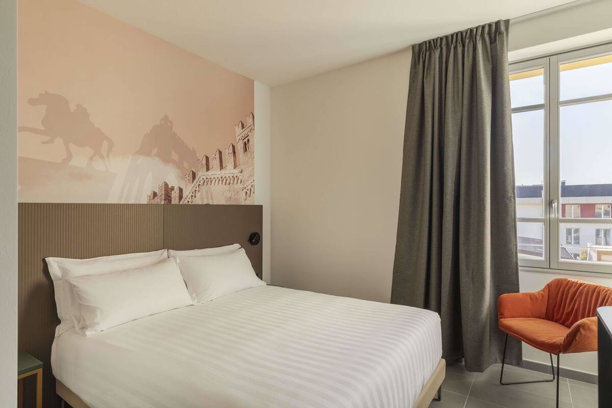Een hotelkamer met een groot bed, een stoel, een muurschildering en een raam met gordijnen.