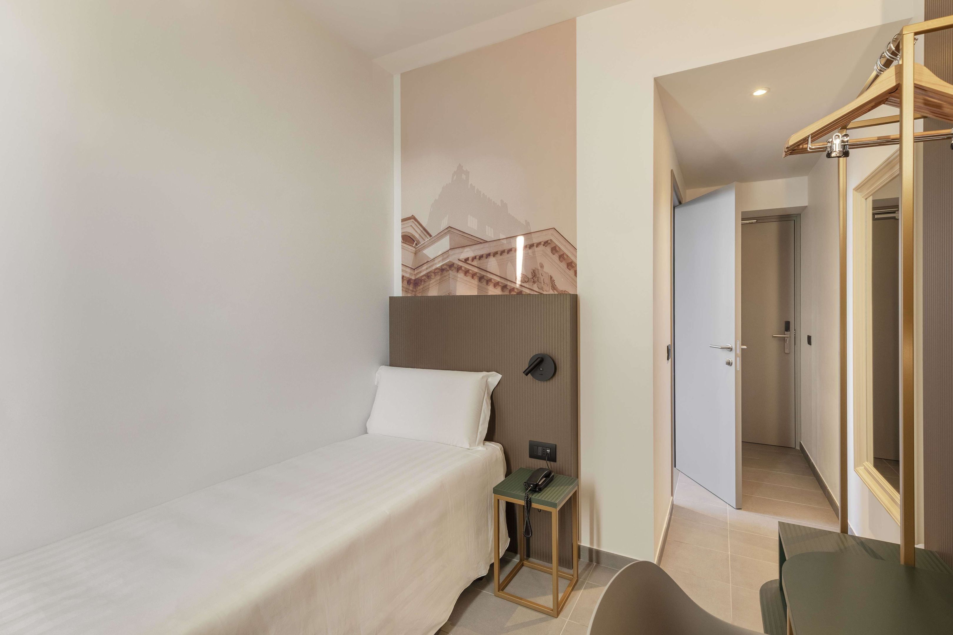 A hotel room with a bed, a small table with a telephone, a coat rack, and a wall-mounted light.