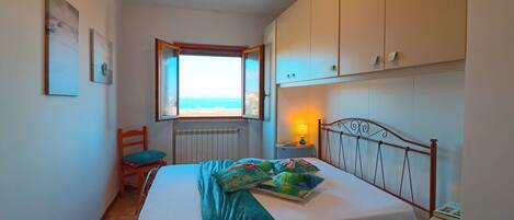 Un dormitorio con cama, una silla, ventana con vistas y mesita de noche con lámpara.