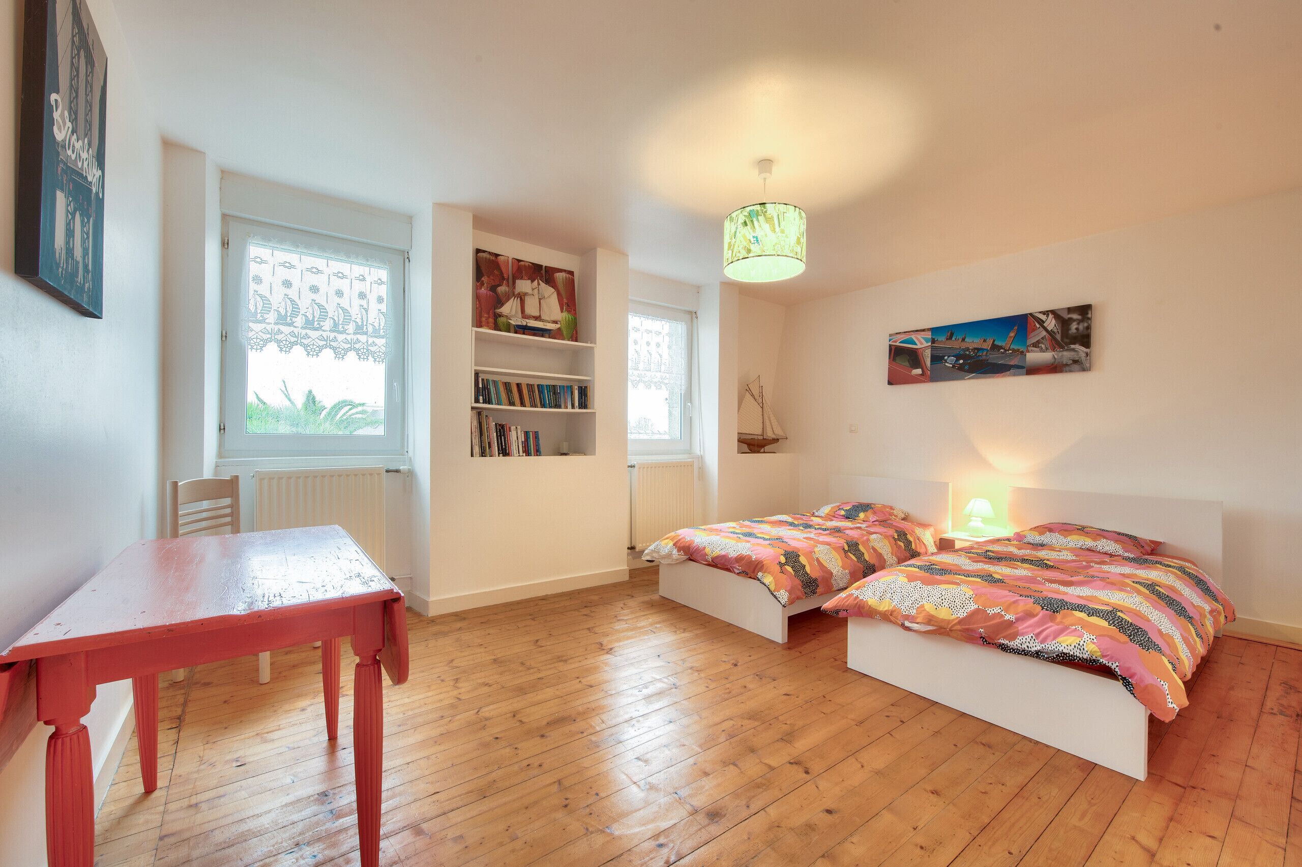 Ein Schlafzimmer mit Holzboden, einem roten Tisch, einem Bücherregal, zwei Betten mit bunter Bettwäsche und einer Hängeleuchte.