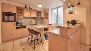 Una cuina moderna amb armaris de fusta, una illa central, tamborets de bar i un gran finestral que proporciona llum natural.