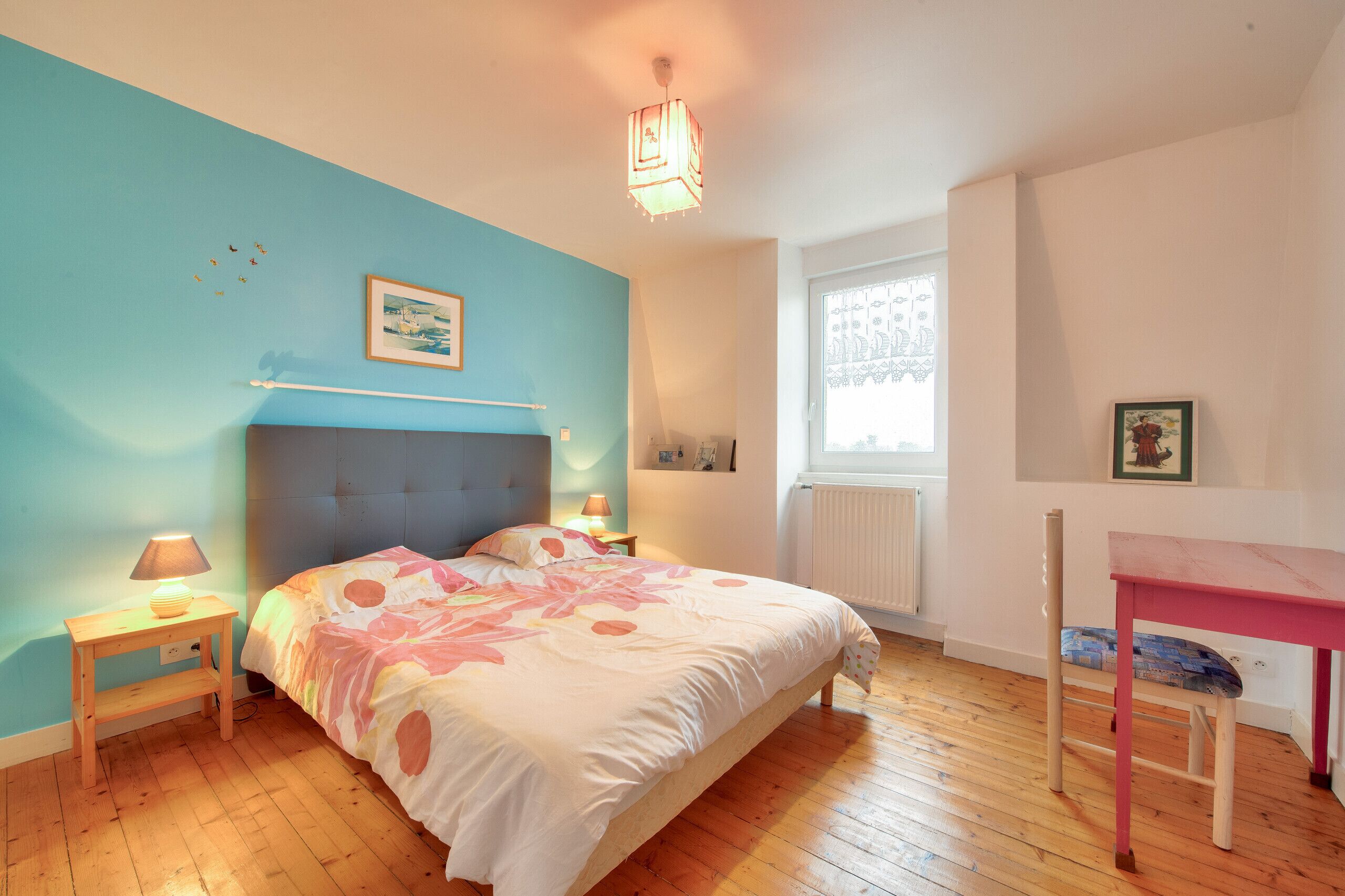 Un dormitorio con cama, mesita de noche con lámpara, ventana y una silla.