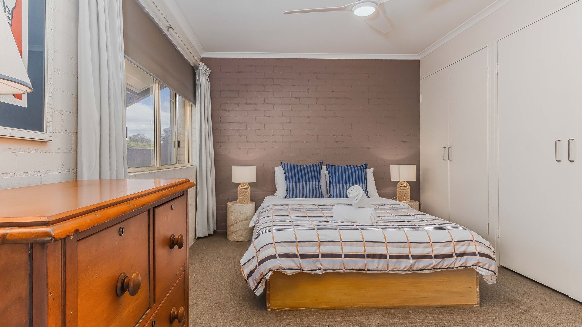 A bedroom with a bed, a wooden dresser, a brick wall, and a ceiling fan.