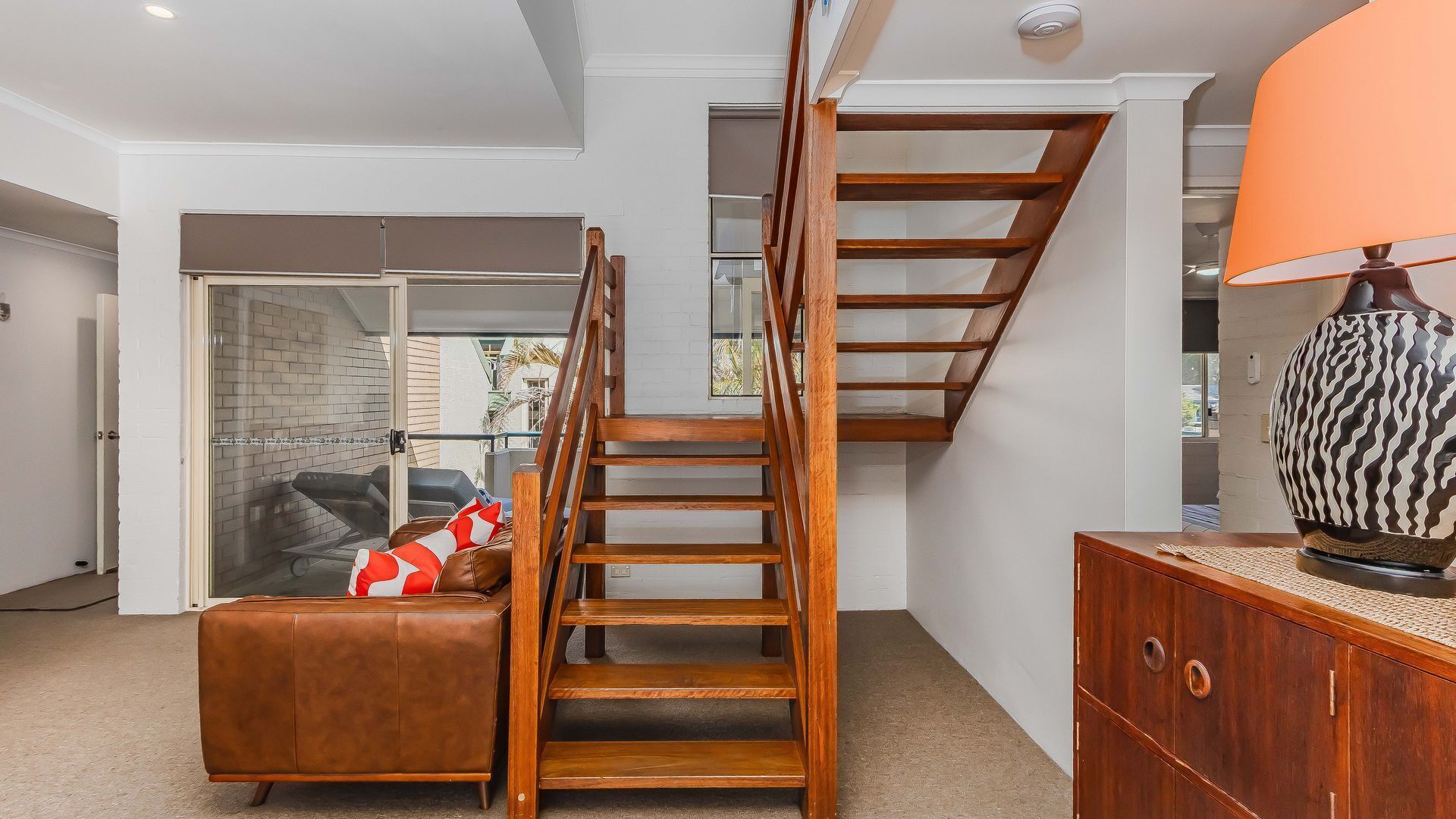 Un escalier avec des éléments en bois, un canapé en cuir agrémenté d’un coussin rouge et blanc, et un meuble en bois surmonté d’un vase décoratif.