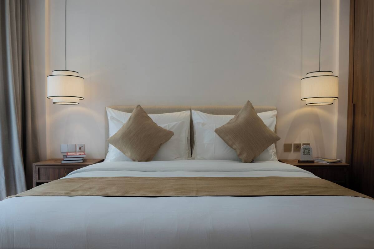 Una cama bien hecha con sábanas blancas y dos almohadas color beige, con dos mesitas de noche a cada lado, cada una con una lámpara.