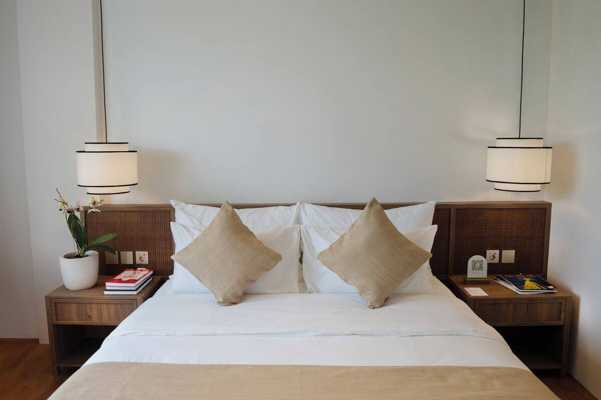 Una cama bien hecha con dos almohadas, una mesita de noche con un libro y una planta, y una lámpara de pared.