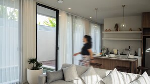 Ein modernes Wohnzimmer mit einer Couch, einem Essbereich und einer Küche im Hintergrund sowie einer Glastür zum Außenbereich.