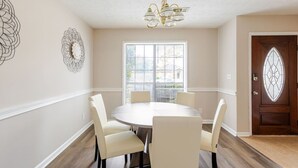 Comedor con una mesa redonda y seis sillas, un candelabro, una ventana con persianas y una puerta decorativa.