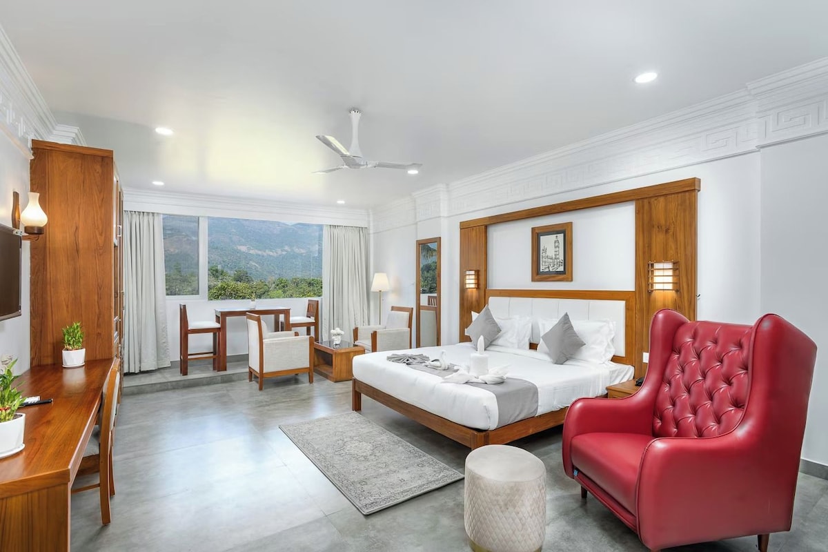 Un cuarto de hotel con cama, un sillón rojo, un escritorio con lámpara y vistas al exterior.