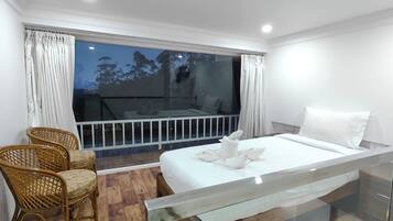 Habitación de hotel con cama, una pequeña zona de estar con sillas, balcón con barandilla y vista a árboles.
