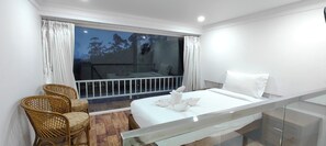 Habitación de hotel con cama, una pequeña zona de estar con sillas, balcón con barandilla y vista a árboles.