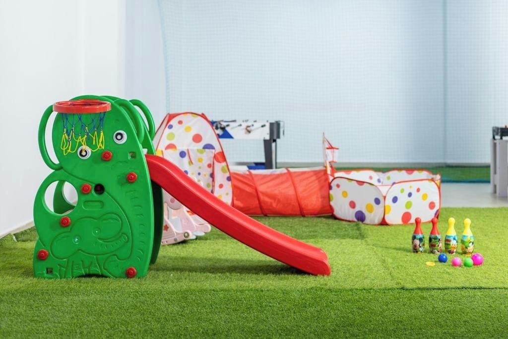 Un área de juegos infantiles con una estructura verde para escalar, un tobogán rojo y una carpa de lunares.