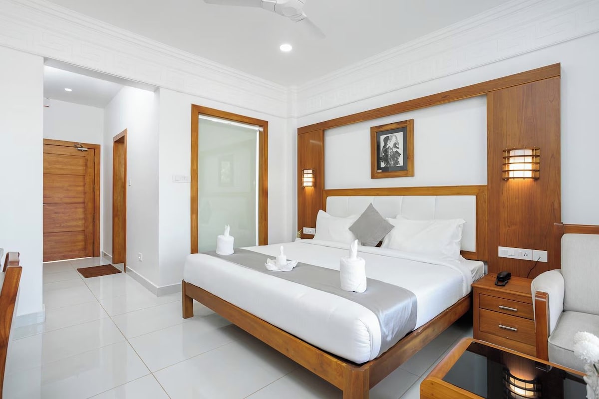 Una habitación de hotel con un marco de cama de madera, ropa de cama blanca, una mesita de noche con una lámpara y otra mesita pequeña con un teléfono.