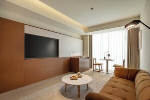 Een moderne woonkamer met een bank, een rond bijzettafeltje met fruit, een flatscreen-tv aan de muur, een klein keukenhoekje en een eettafel met stoelen.