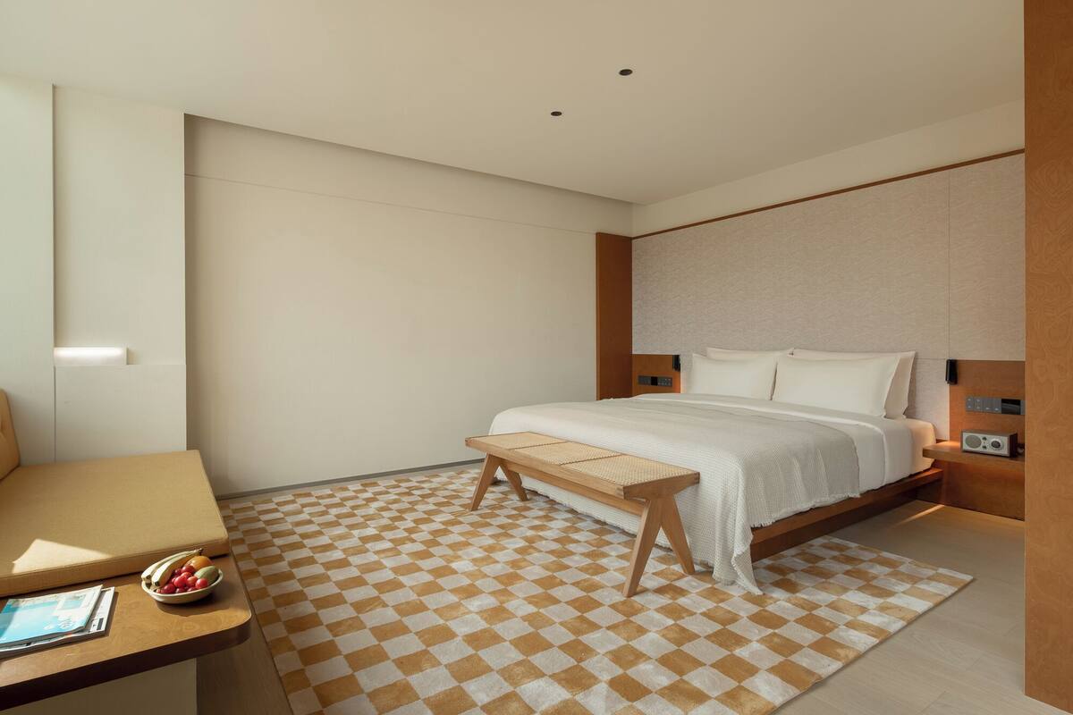 Una habitación de hotel moderna con suelo a cuadros, una cama con sábanas blancas, un banco de madera y una mesita de noche con frutero.
